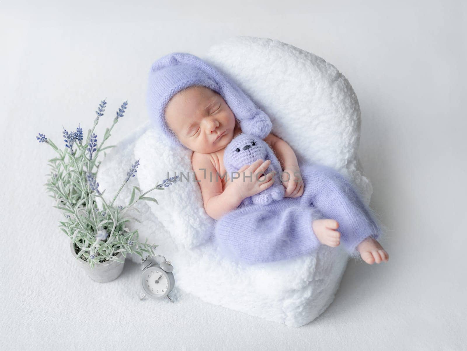 Newborn Baby In Lilac Pants And Cap Sleeps In Tiny Chair During Studio Photoshoot