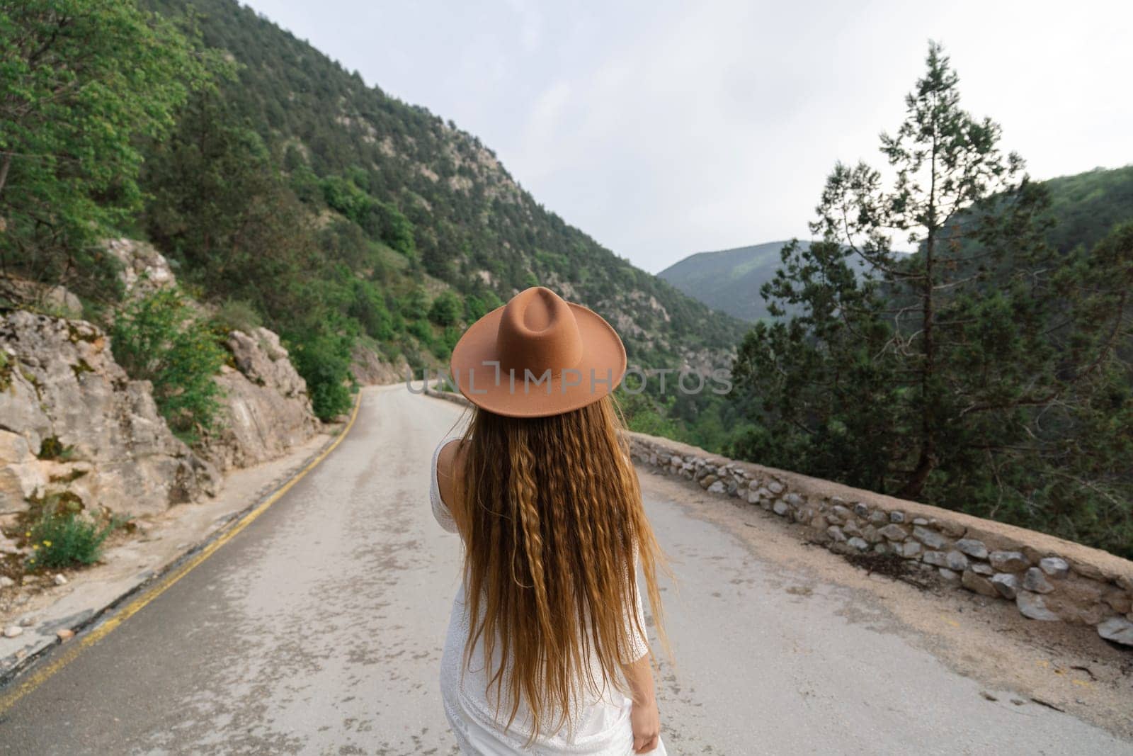 A woman wearing a brown hat walks down a road. The road is empty and the sky is cloudy