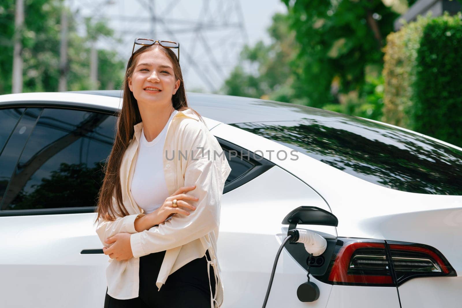 Young woman recharge her EV electric vehicle at green city park parking lot. Urban sustainability lifestyle for environmental friendly EV car with battery charging station. Expedient