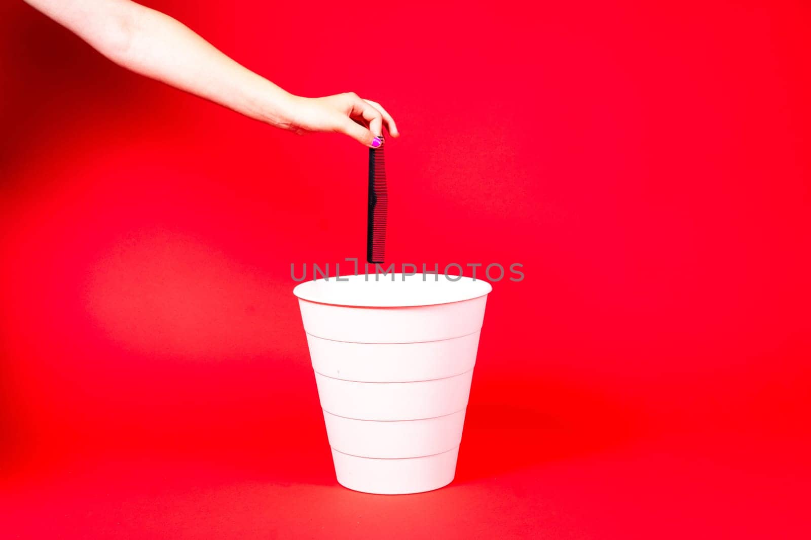 Hand throws plastic comb into trash bin with package on red background by Zelenin