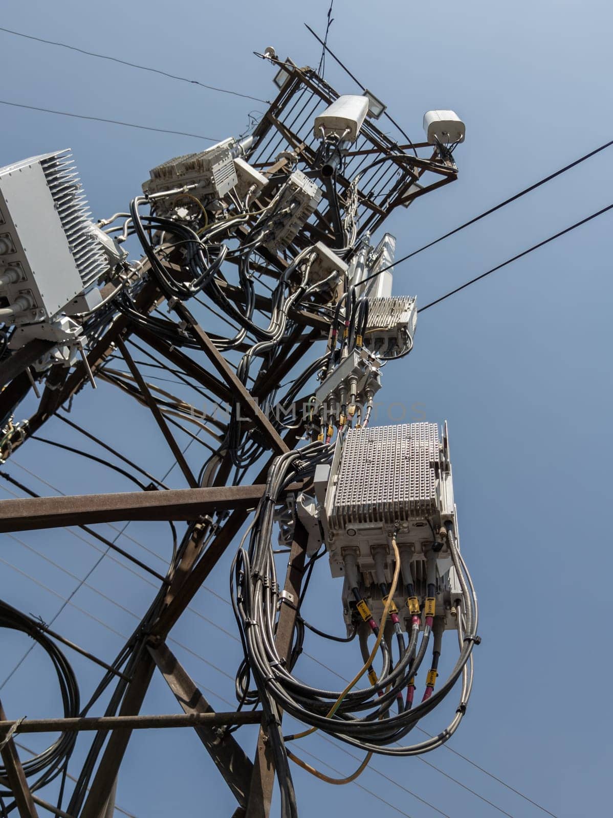 Detailed view of a cluttered utility pole, featuring an assortment of electronic equipment, including multiple antennas, circuit boxes, and tangled wires against a clear blue sky.
