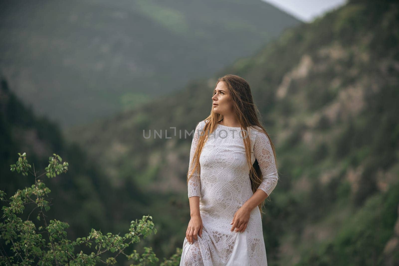 A woman in a white dress stands on a mountain top, looking out over the landscape. The scene is serene and peaceful, with the woman's long hair blowing in the wind