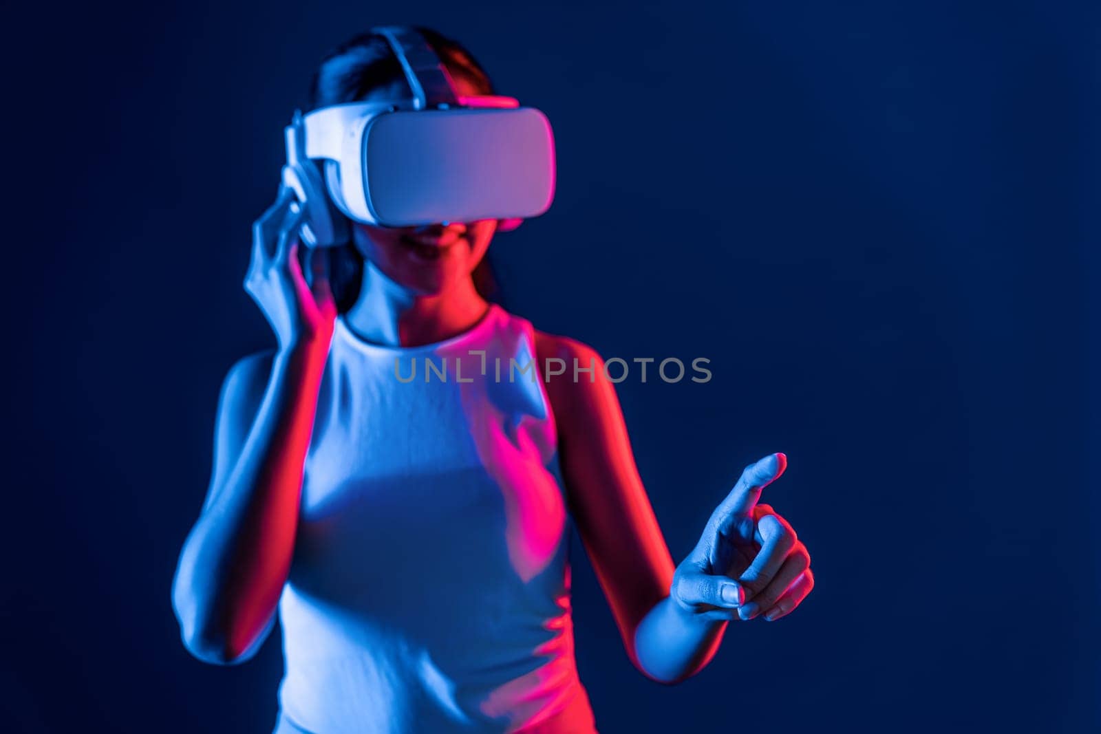 Smart female stand with surrounded by cyberpunk neon light wear VR headset connecting metaverse, futuristic cyberspace community technology. Woman using finger pointing virtual object. Hallucination.