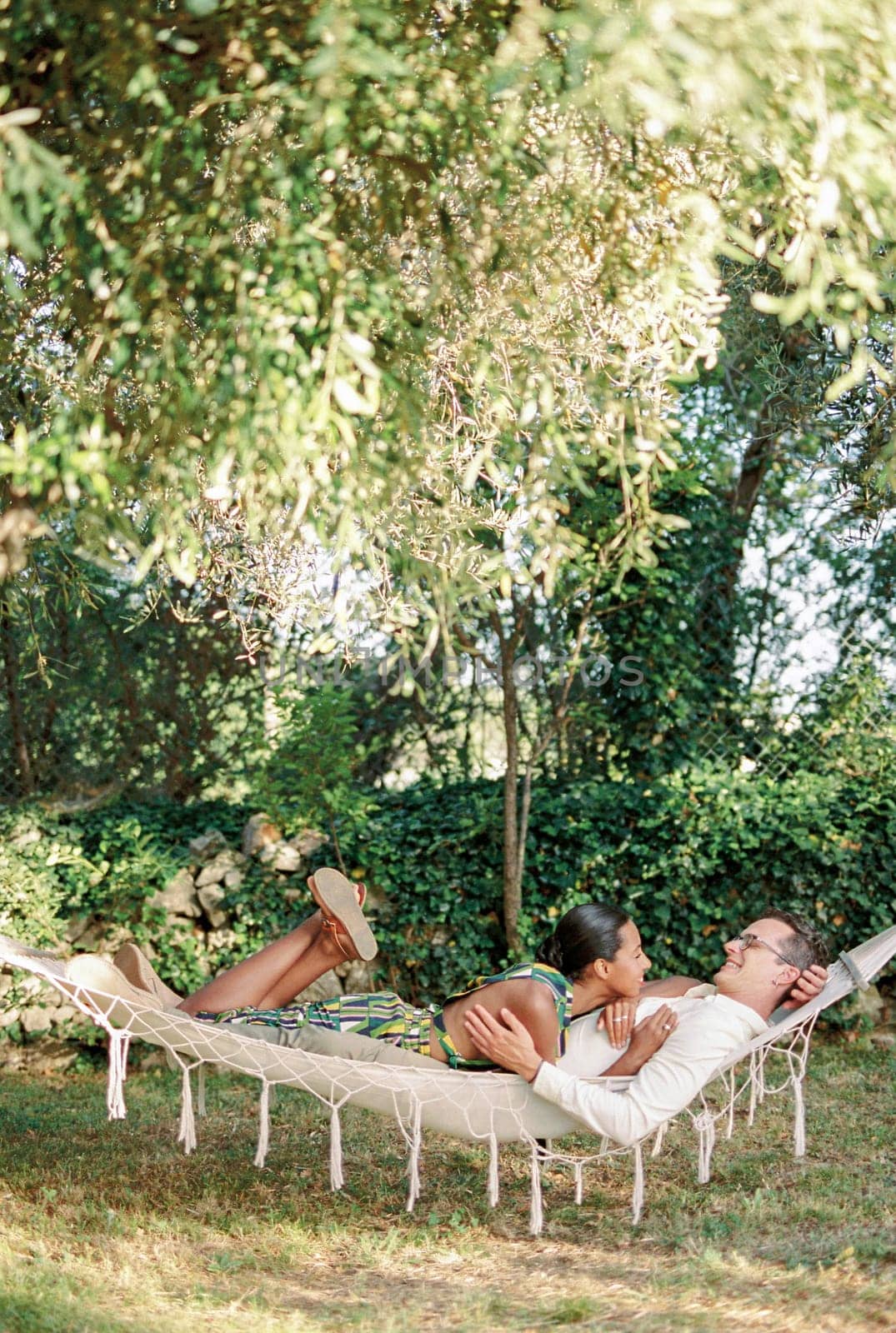 Woman resting her head on her arm on the chest of man hugging her waist while lying in a hammock in the garden. High quality photo
