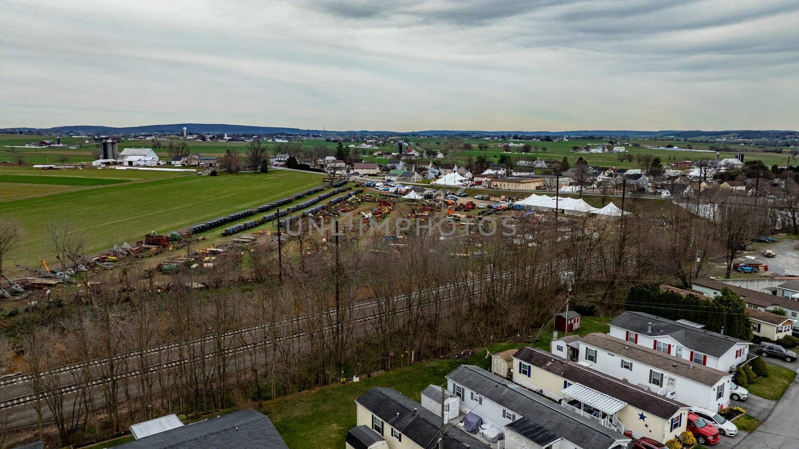 A drone photo capturing a lively rural community with residential areas, agricultural machinery, and a small fair setup under an overcast sky.