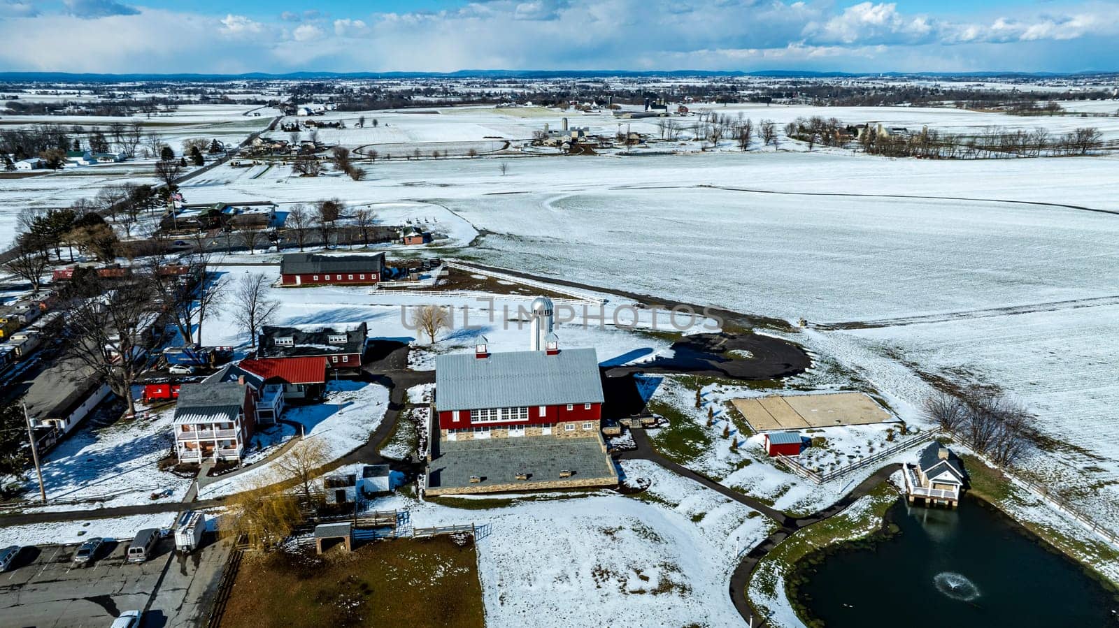 An Aerial View Of A Snowy Rural Community With A Central Pond And Surrounding Buildings.