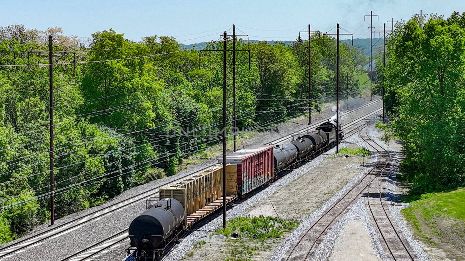 An engaging aerial view of a freight train carrying diverse cargo winding through rural tracks surrounded by lush greenery, under a bright sunny sky.