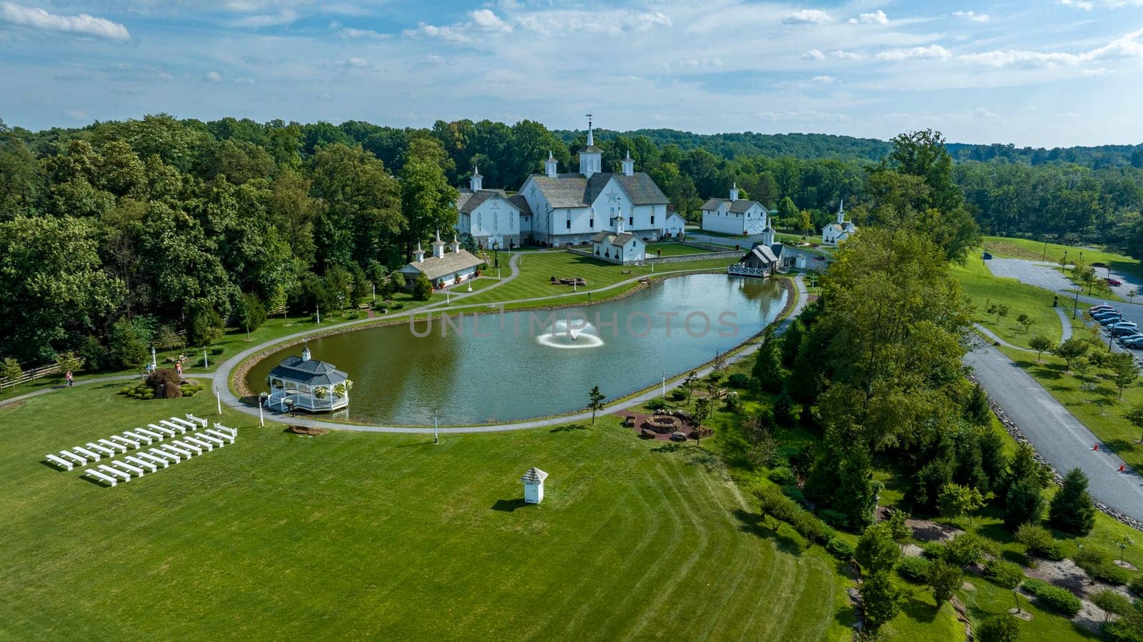 Aerial View Showcasing A Cluster Of Traditional White Orthodox Churches With Cross-Topped Domes, Arranged Around A Curved Pond With A Fountain, Amidst Green Trees And Neatly Arranged White Benches