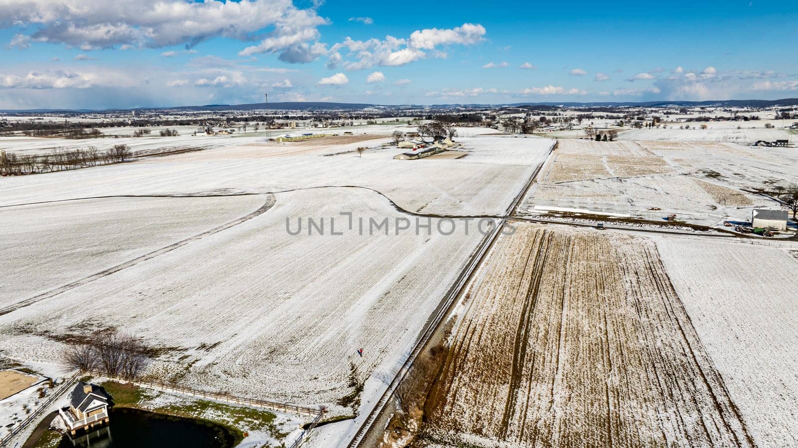 An Aerial View Of Expansive Snow-Dusted Farmland With Rural Buildings And Silos.