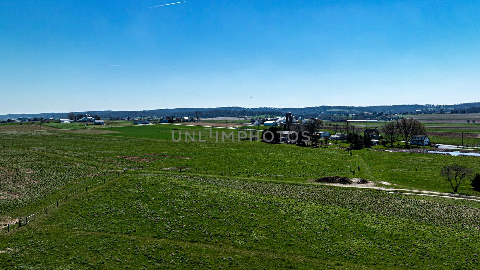 An expansive view from above showcasing lush green pastures segmented by fencing, with a detailed farmstead and distant hills under a brilliant blue sky.