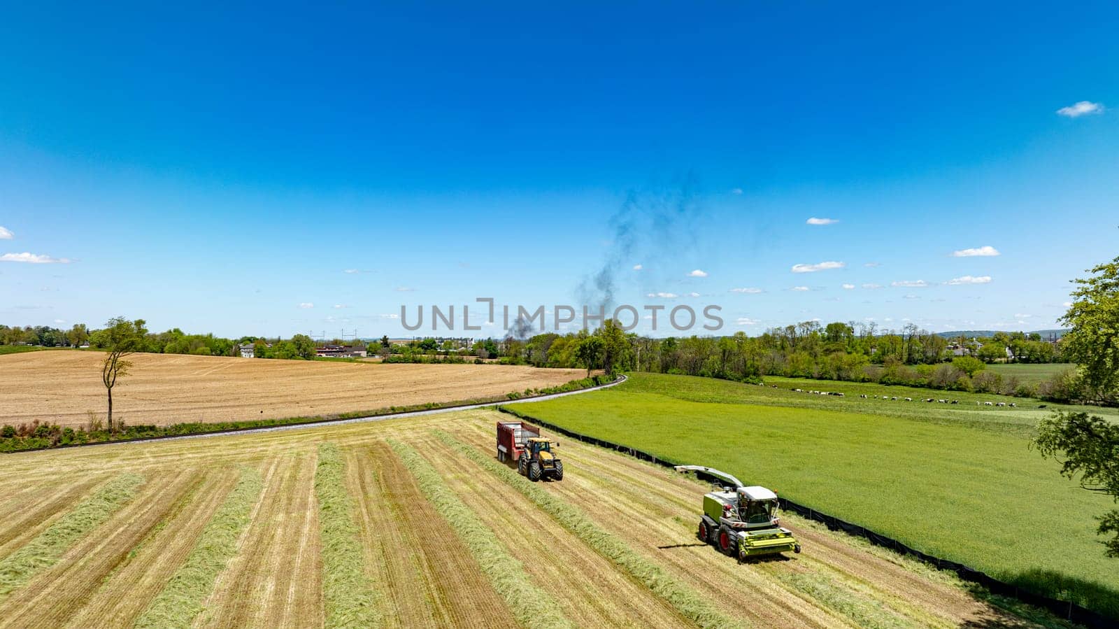 An aerial shot displaying intense harvesting activity with machinery in action across a multi-textured landscape of cut and uncut fields, under a bright blue sky with distant smoke visible.