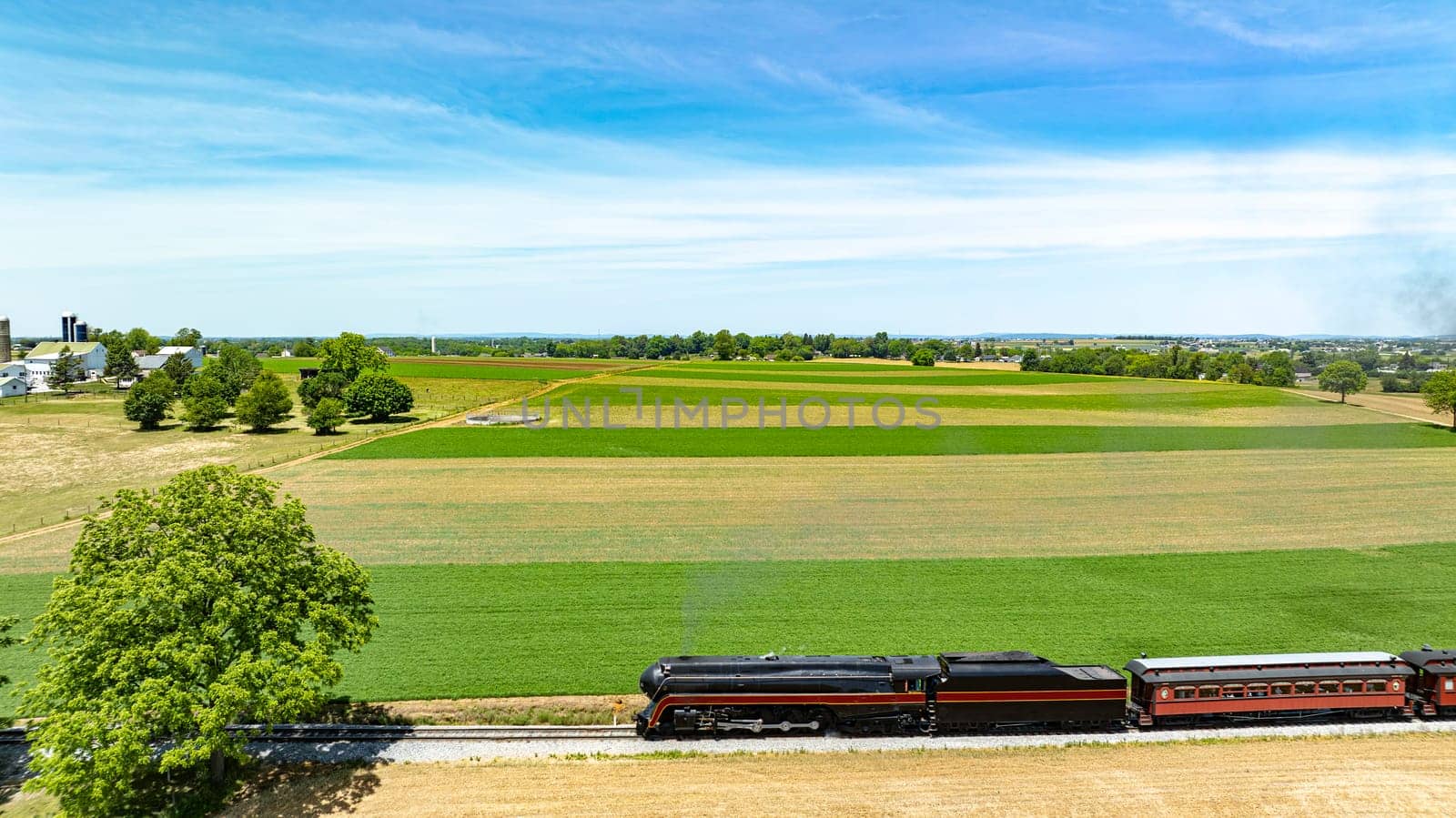 Stunning aerial view of a classic train passing through expansive fields, weaving its path between lush greenery and farmland under a clear sky.