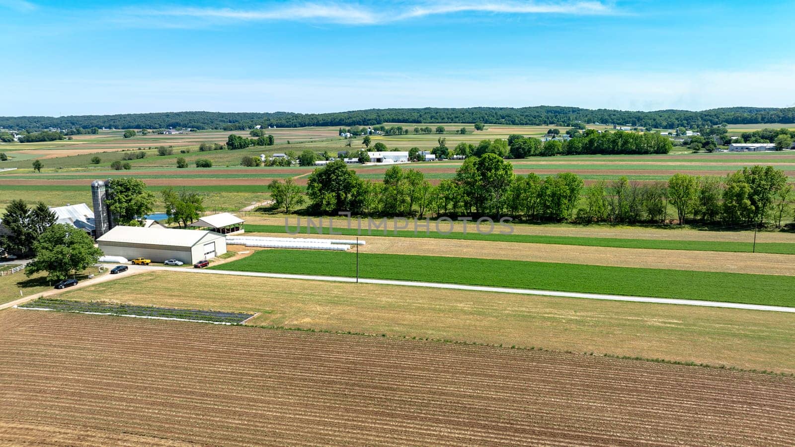 An expansive aerial shot capturing the serene beauty of a rural farm, showcasing a mix of plowed fields, green crops, and farm buildings under a clear blue sky.