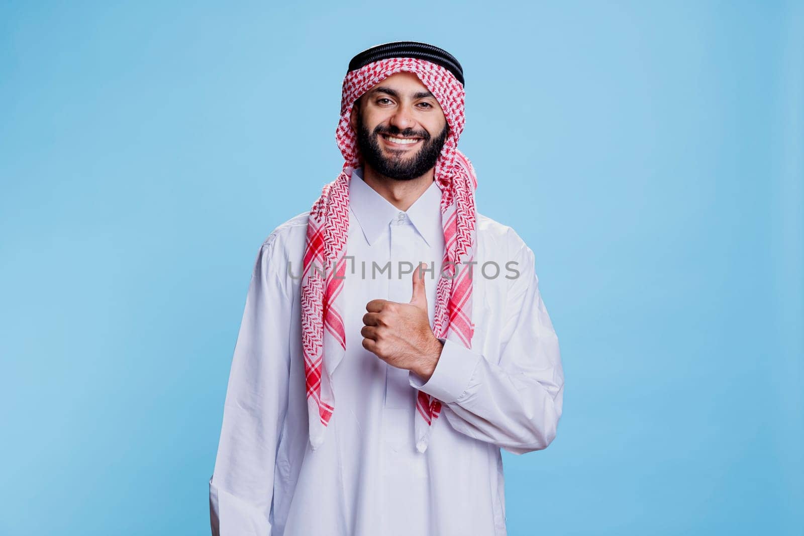 Arab man wearing islamic traditional clothes posing with thumb up signifying approval and agreement portrait. Smiling muslim person in thobe and ghutra showing like gesture