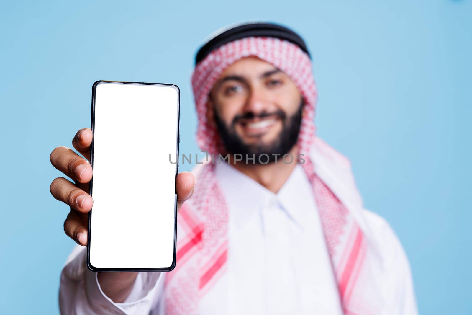 Man in arabic attire holding smartphone displaying white screen for application advertising. Smiling muslim person posing in studio and showing mobile phone blank touchscreen closeup