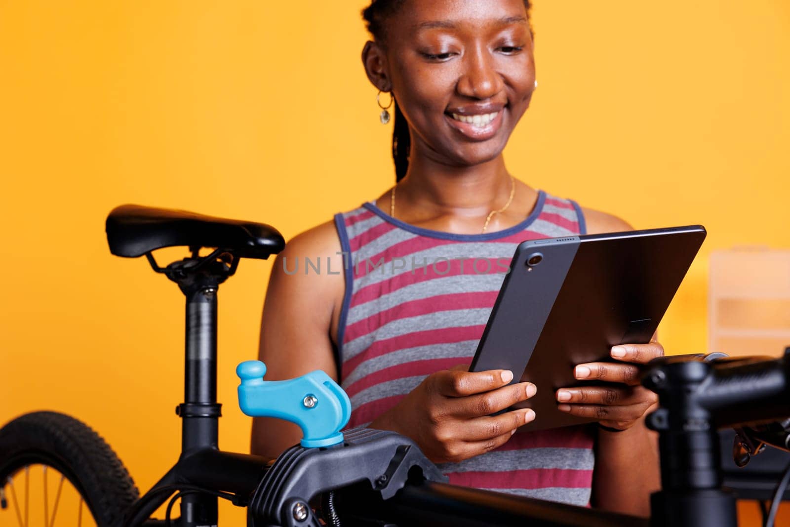 Energetic black woman carefully inspects and repairs bike frame using repair-stand and digital device. Image showing close-up view of female cyclist holding smart tablet while servicing bicycle.