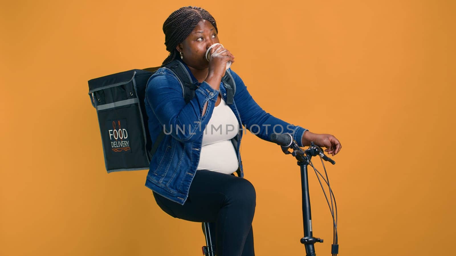 Young courier on bicycle takes relaxing moment to enjoy coffee while delivering packages. African american lady using drink to quench her thirst while providing efficient food delivery service.