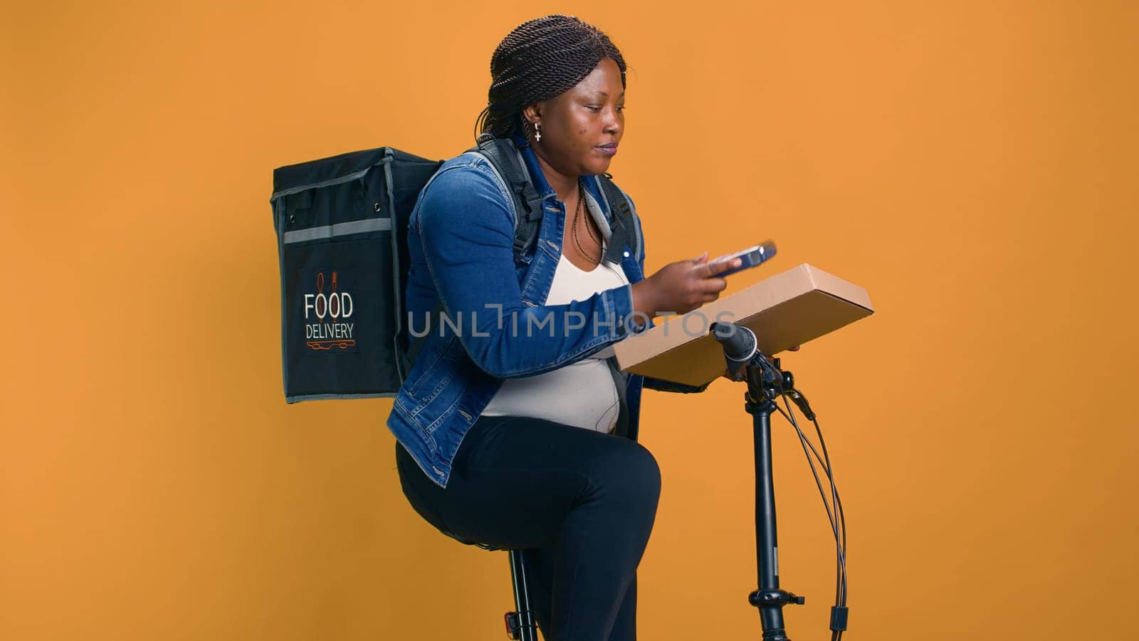 Black woman delivers takeout orders on bicycle, utilizing wireless payment device for seamless transactions. African american delivery person taking pos machine from pocket for contactless pay.