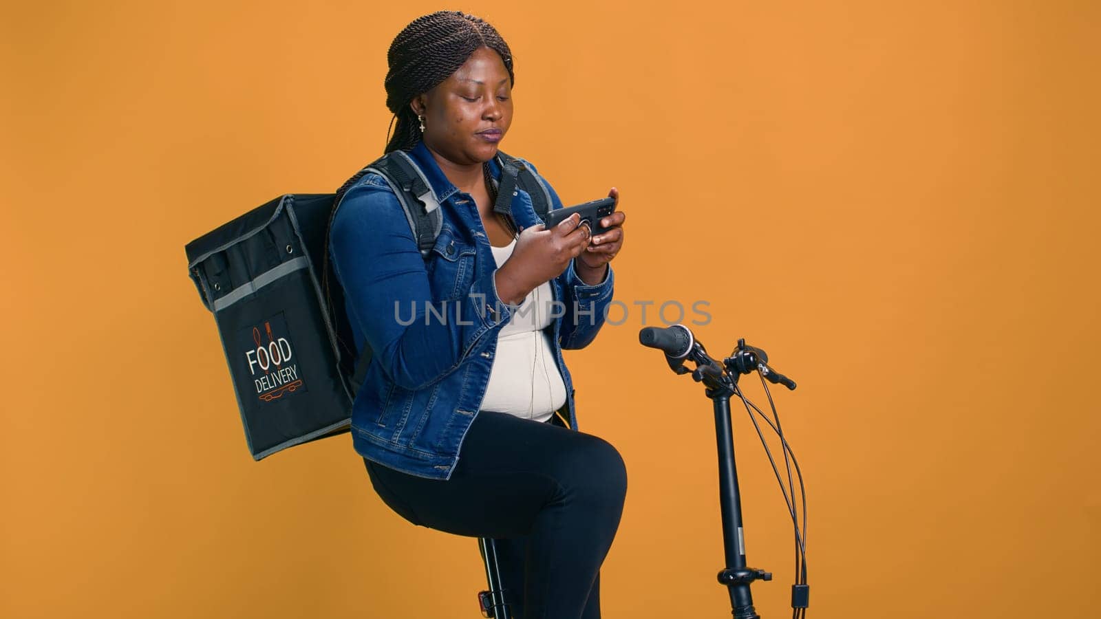 Professional african american cyclist uses bicycle and smartphone to offer efficient food delivery service. Young black woman embraces fast-paced economy of utlizing mobile device for courier job.