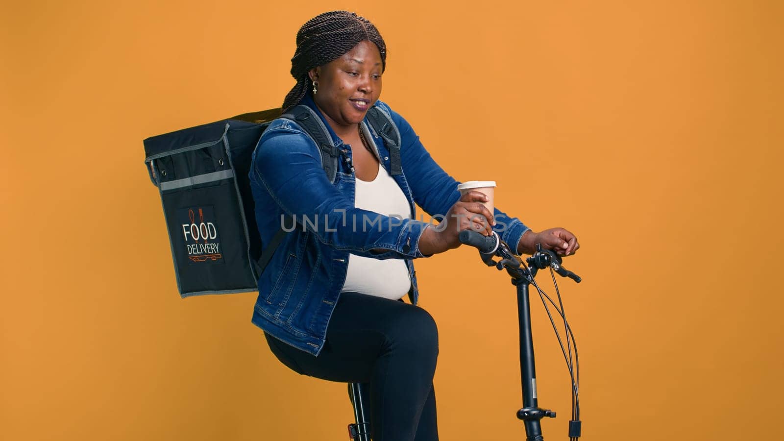 African american delivery woman multitasking carrying coffee in hand and riding bicycle in neighborhood. Active courier on the go having quick energizing refreshment before going for food delivery.