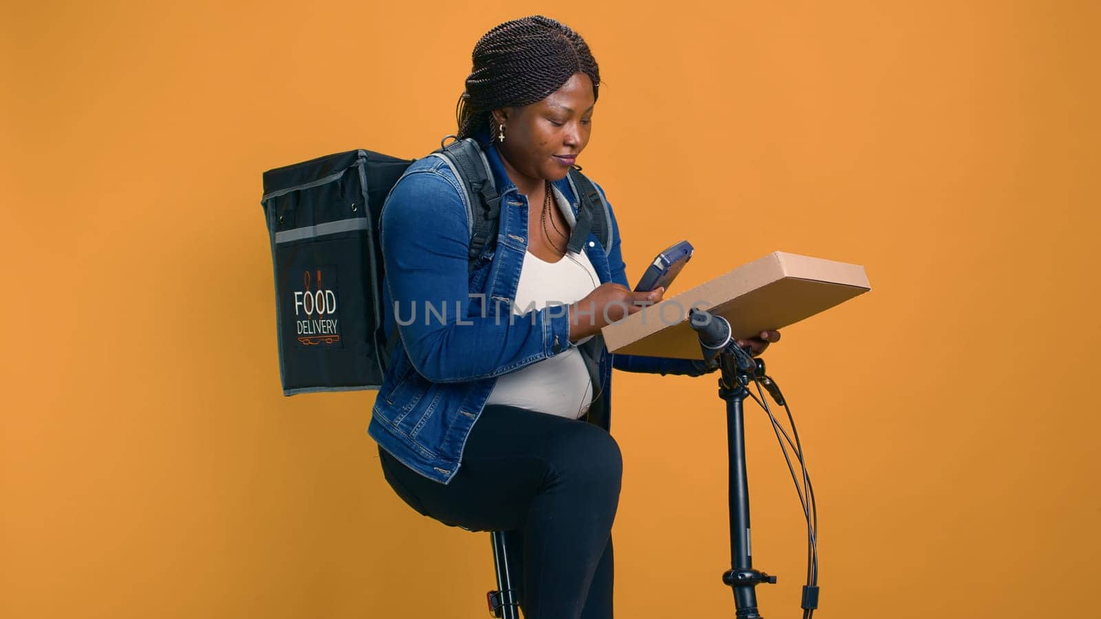 African American courier delivers package using bicycle and modern pos machine ensuring contact-free service. Delivery woman providing nfc terminal for convenient wireless payment for food delivery.