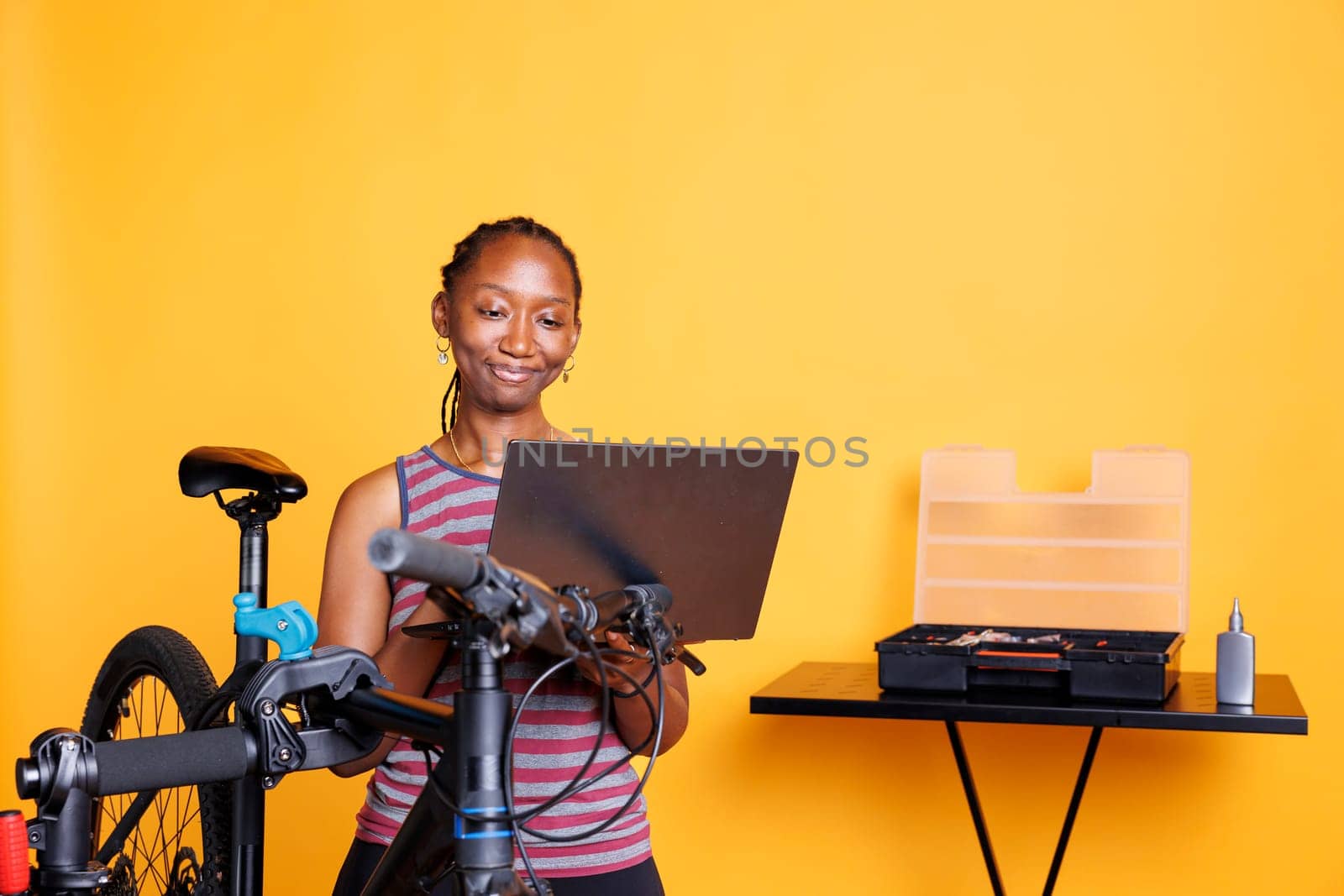 In front of yellow background, black woman performs bike maintenance by utilizing digital laptop for instructions. African american female using minicomputer and professional tools for fixing bicycle.