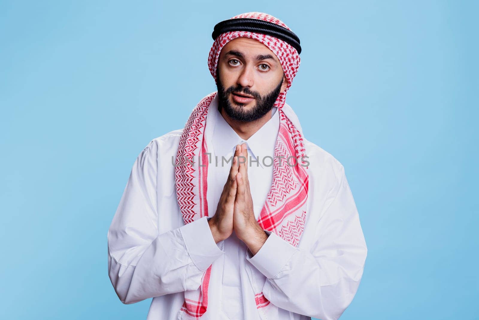 Man wearing traditional muslim thobe and ghutra clothes praying studio portrait. Arab person standing with folded hands showing symbol of faith and spirituality while looking at camera