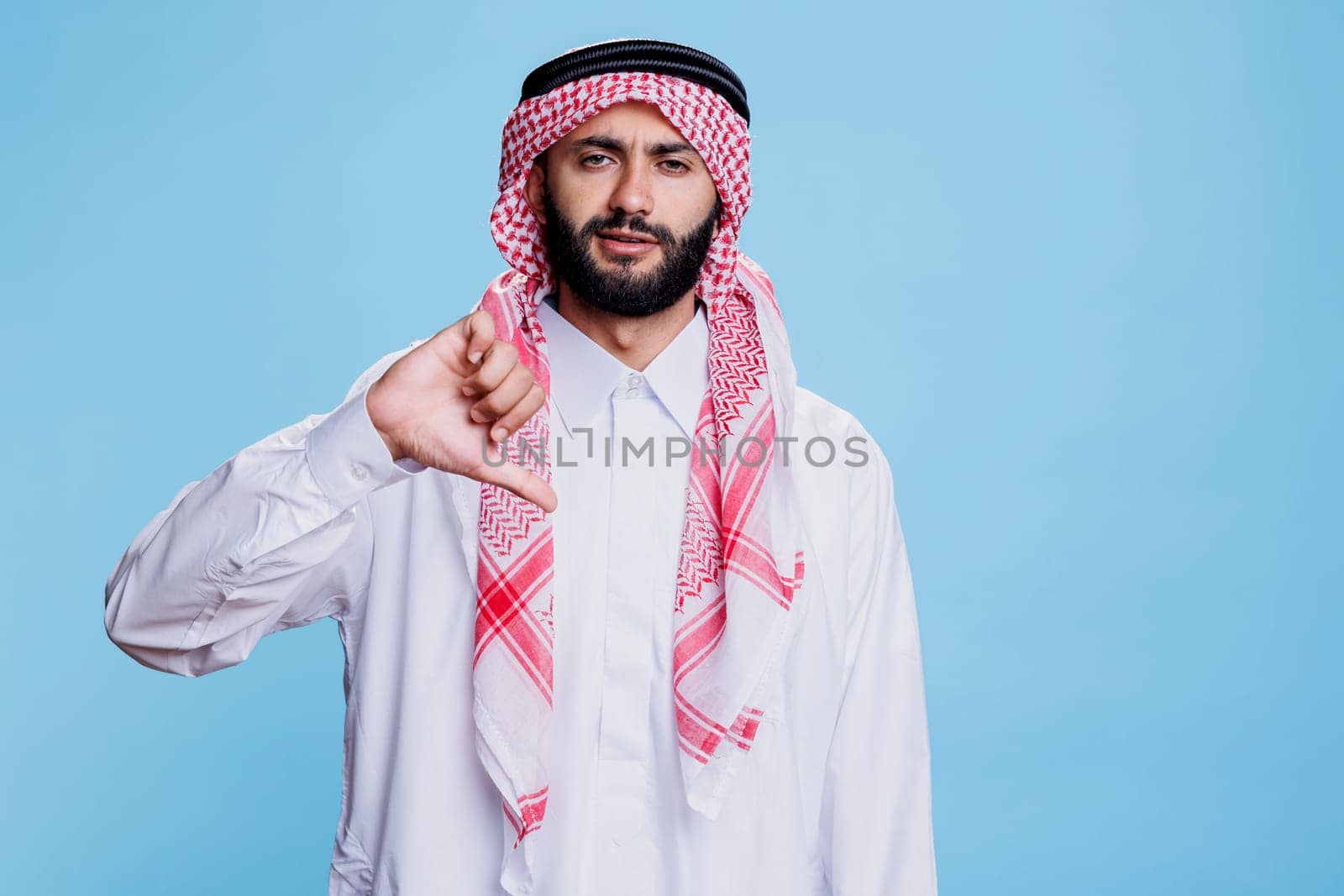 Muslim man dressed in traditional arabian clothes showing thumb down studio portrait. Arab in thobe and ghutra showcasing disagreement and looking at camera with serious expression