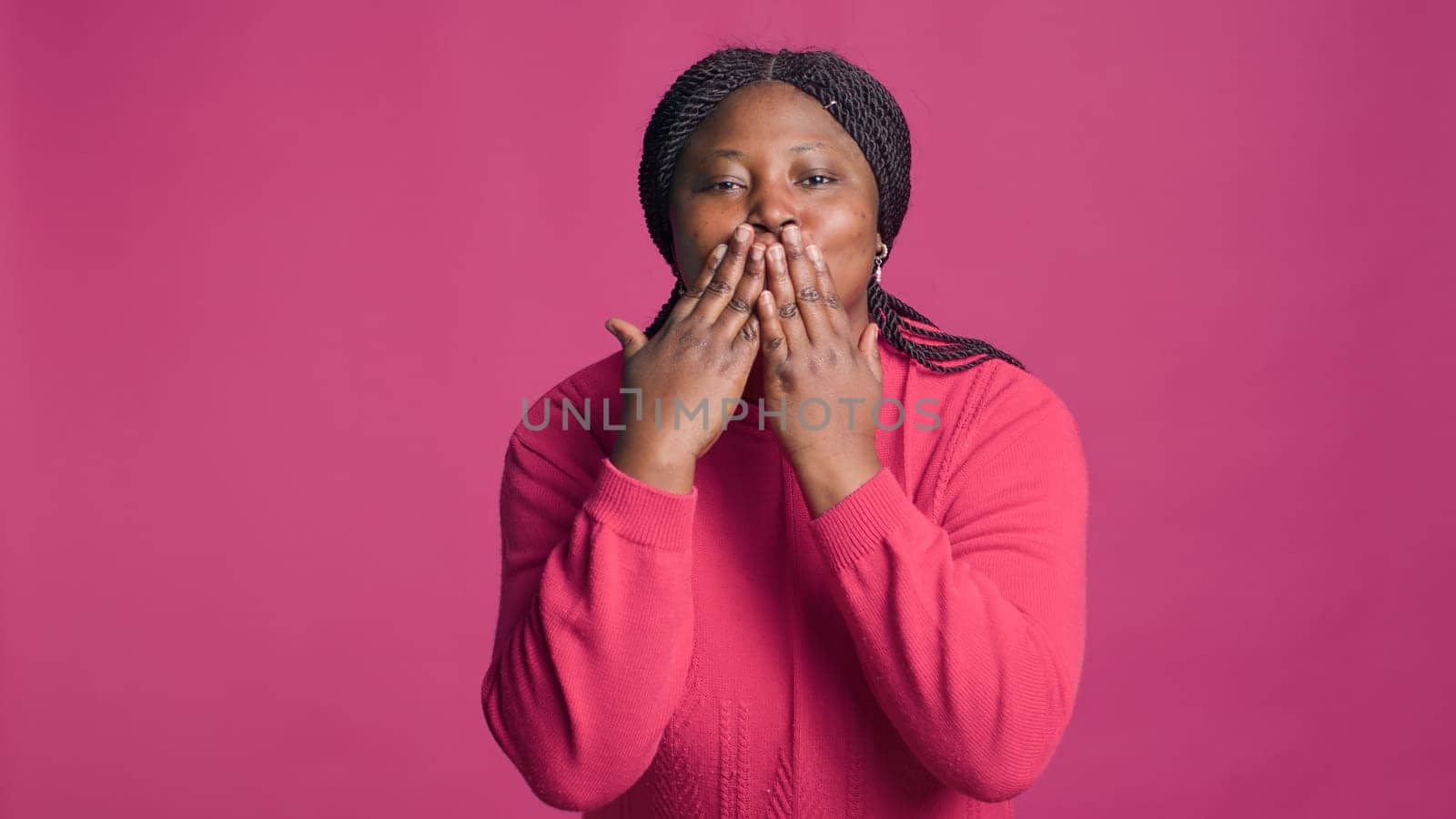 Lady with african american ethnicity blowing a kiss towards camera being lovely isolated on pink background. Stunning joyful black woman expressing love with cute kissing hand gestures.