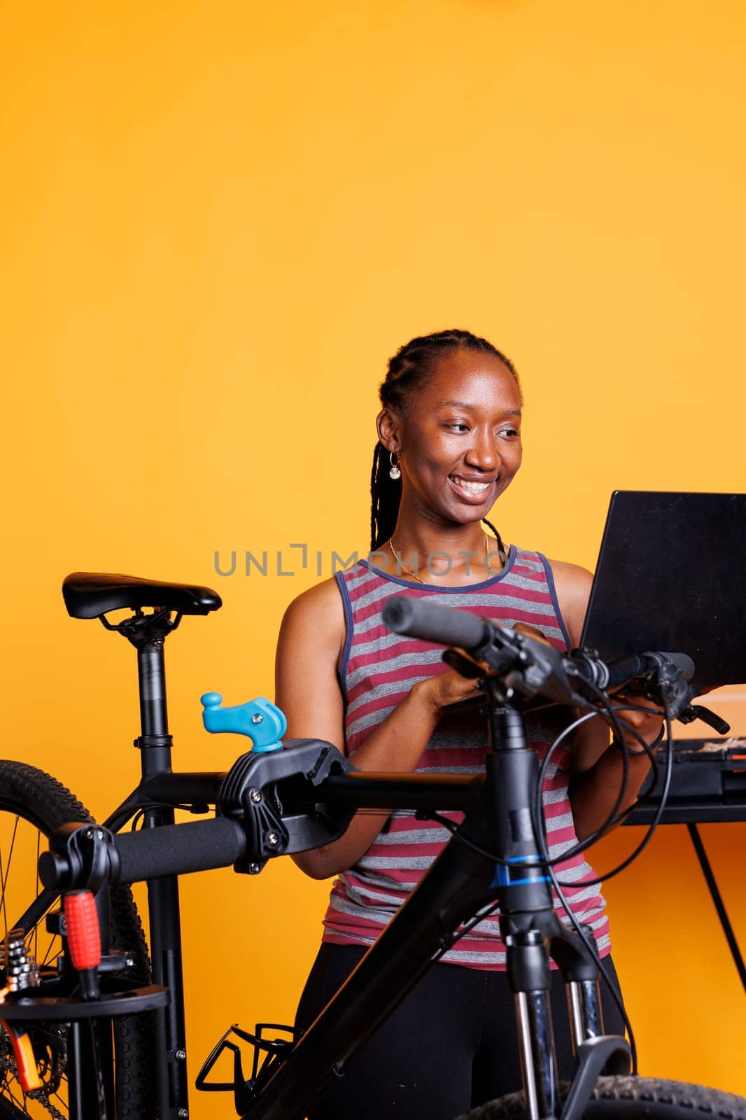 Young African American female repairs damaged bicycle using toolkit and wireless computer. Active vibrant black woman surfing the internet for bike maintenance, in front of isolated yellow background.
