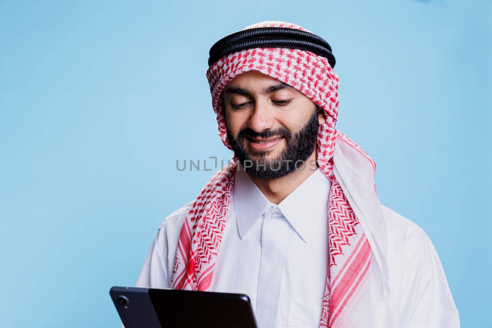 Smiling man wearing arabic clothes using digital tablet and scrolling social media. Cheerful muslim person dressed in ghutra headscarf and thobe tapping on portable device screen