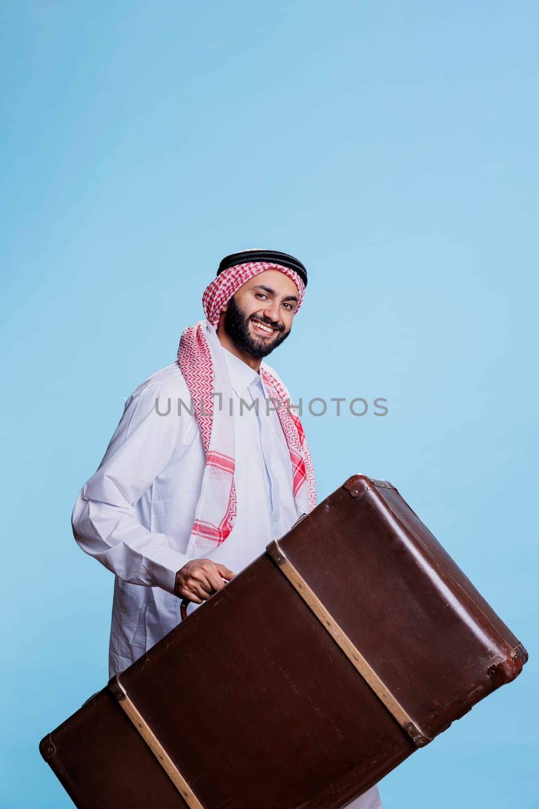 Smiling man wearing cultural muslim clothes carrying vintage suitcase while traveling studio portrait. Cheerful arab holding luggage while going on trip and looking at camera