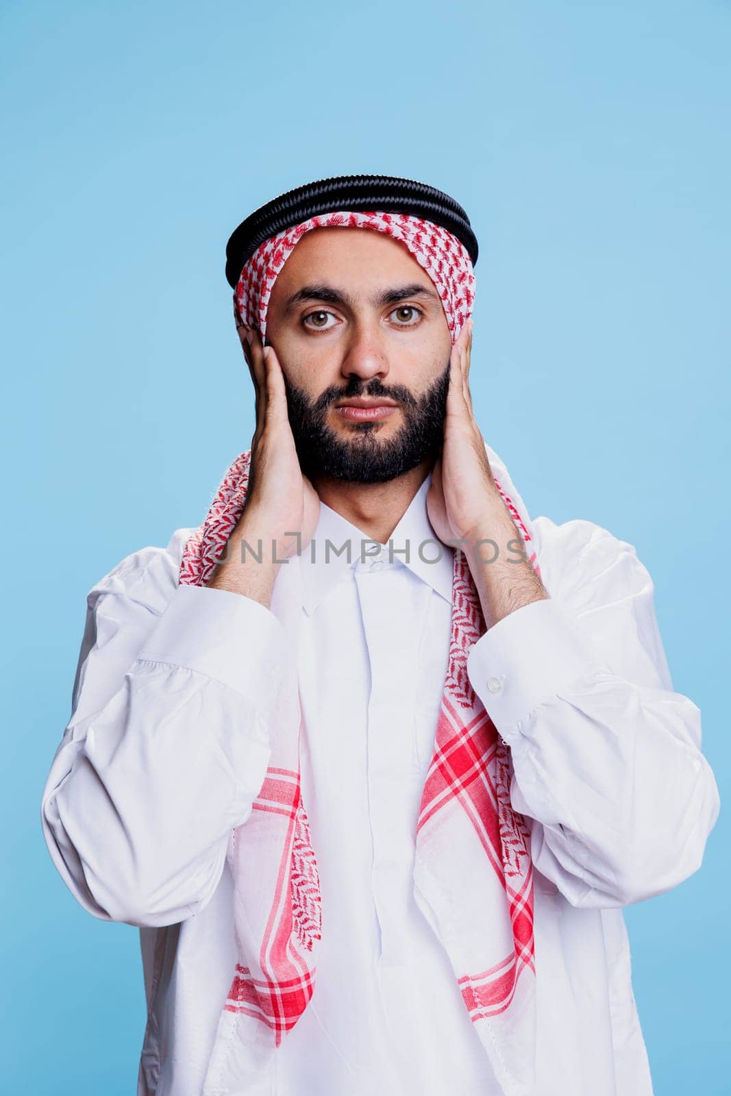 Man wearing traditional muslim clothes covering ears with hands while showing hear no evil three wise monkeys concept. Arab person ignoring loud sound and looking at camera