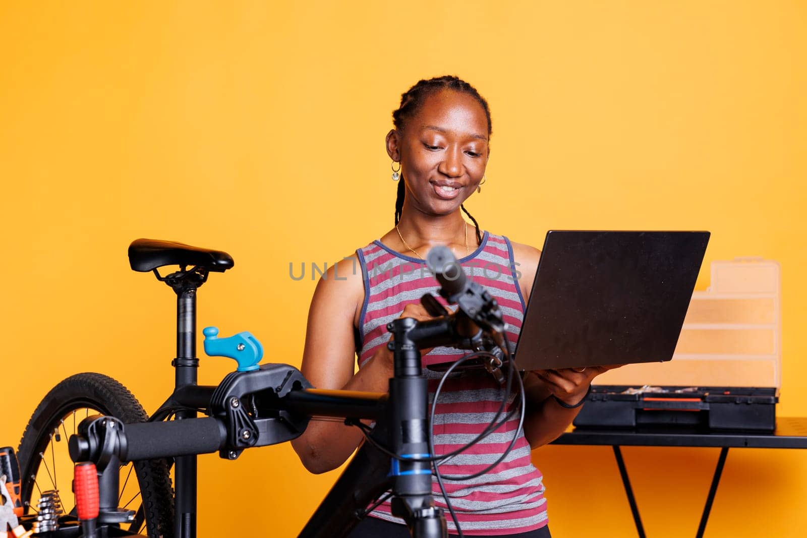 Sporty african american lady repairing broken bicycle with assistance from internet on digital laptop. Youthful black woman examining and fixing damaged components with toolkit and personal computer.