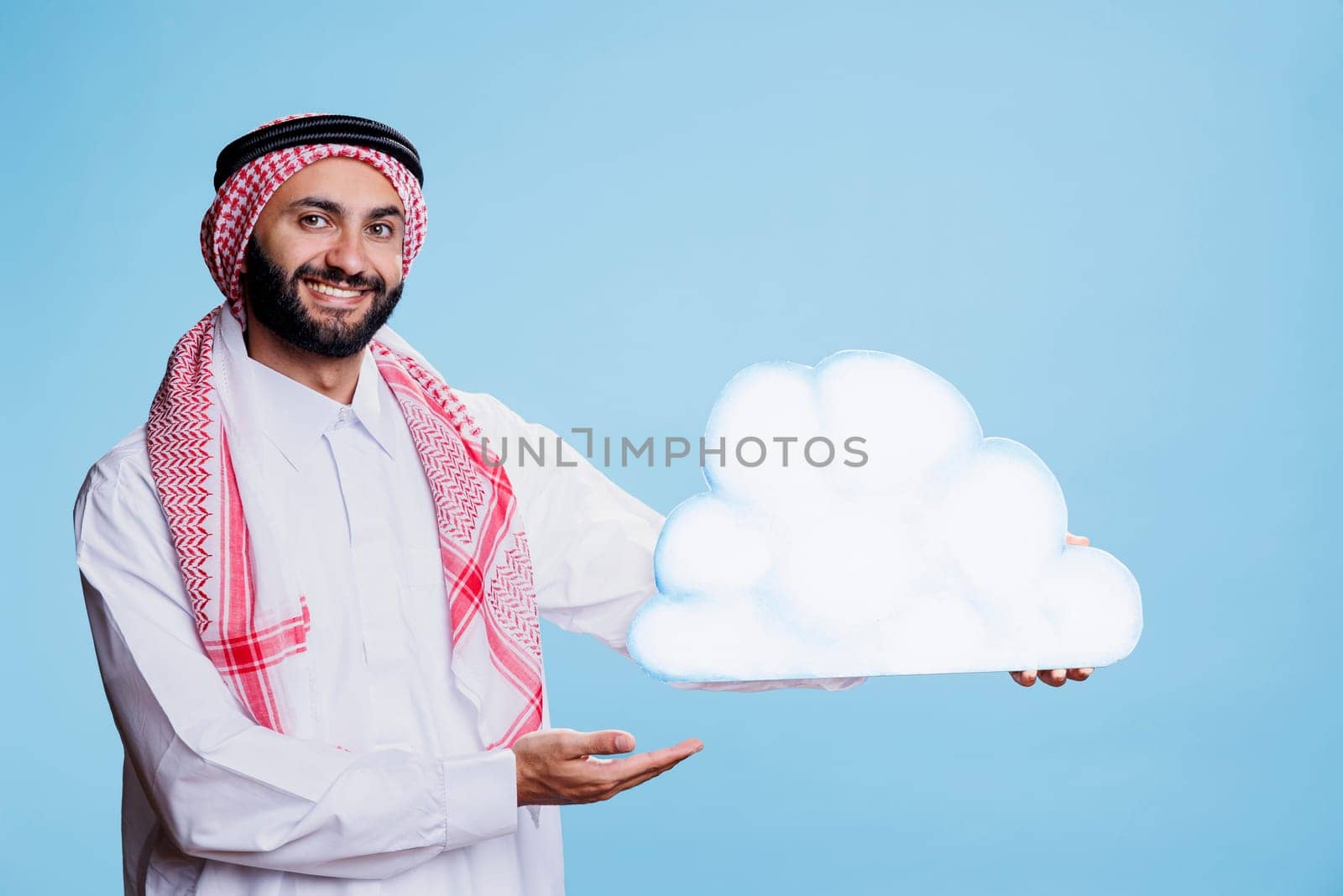 Carefree man wearing arabic traditional clothes posing with cloud shape cardboard banner studio portrait. Smiling muslim person holding showing white bubble and looking at camera