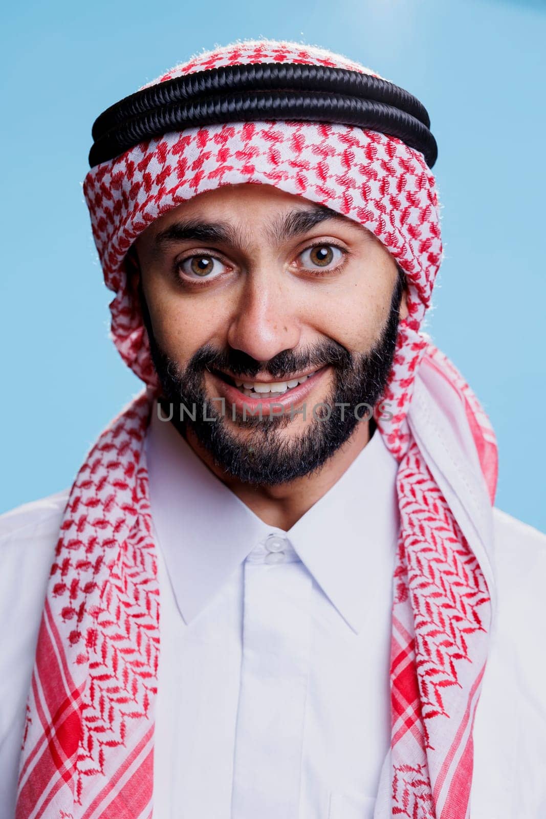 Excited muslim man wearing traditional islamic thobe and headscarf looking at camera with carefree expression. Cheerful smiling arab dressed in ghutra headdress closeup portrait