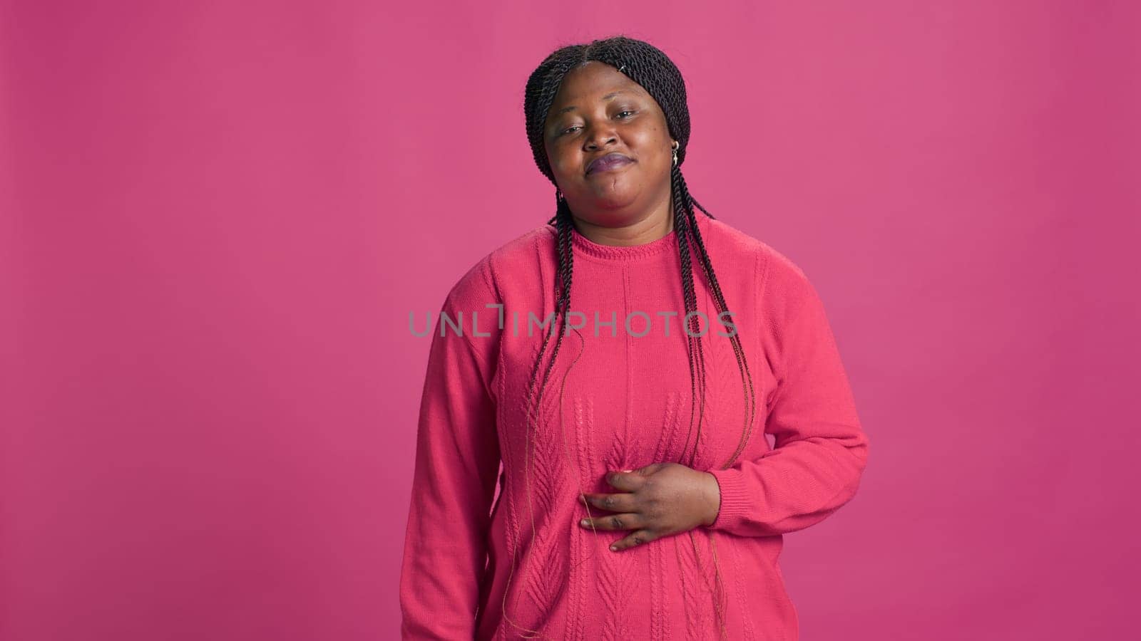 Youthful female looking at camera and showing unhappy facial expression against pink backdrop. Sorrowful african american woman standing in front of isolated background.