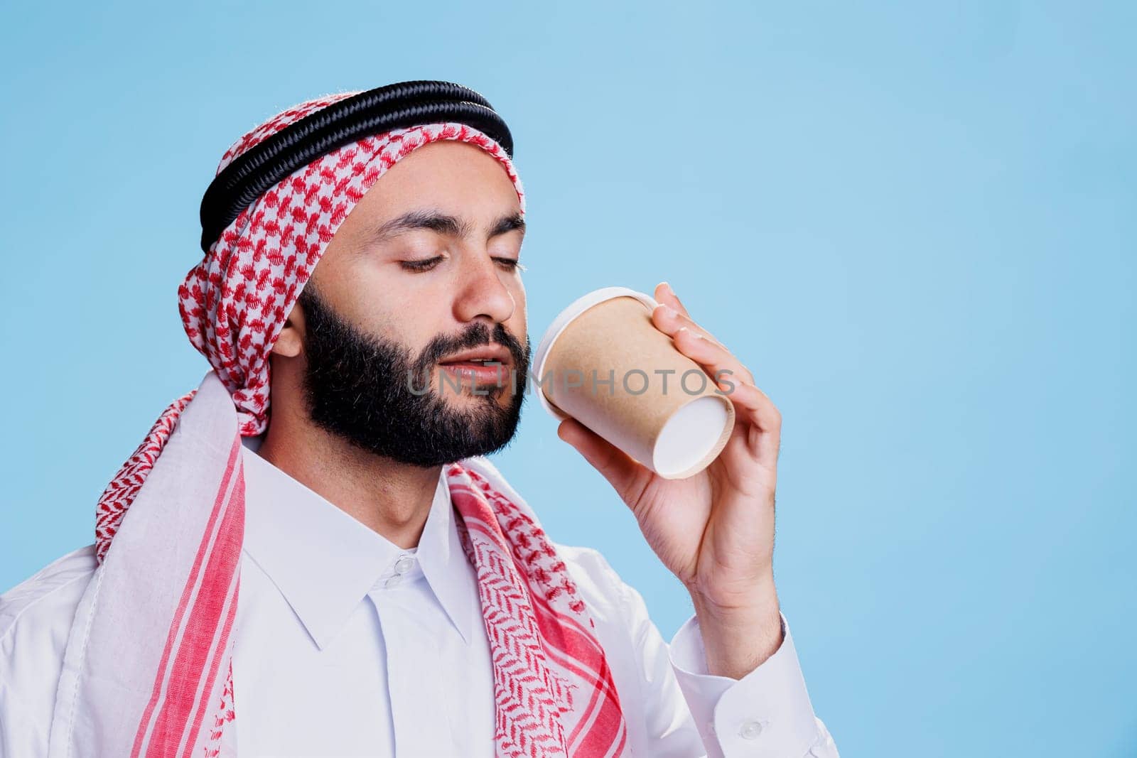 Muslim man wearing traditional islamic clothes drinking coffee to go from paper cup. Arab person dressed in thobe and headscarf holding mug and enjoying hot takeaway beverage