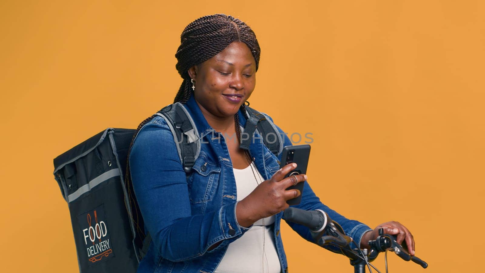Active black woman delivering food on bicycle with cell phone in hand. African american female checking delivery app on smartphone for transportation of package. Portrait shot, smiling at camera.