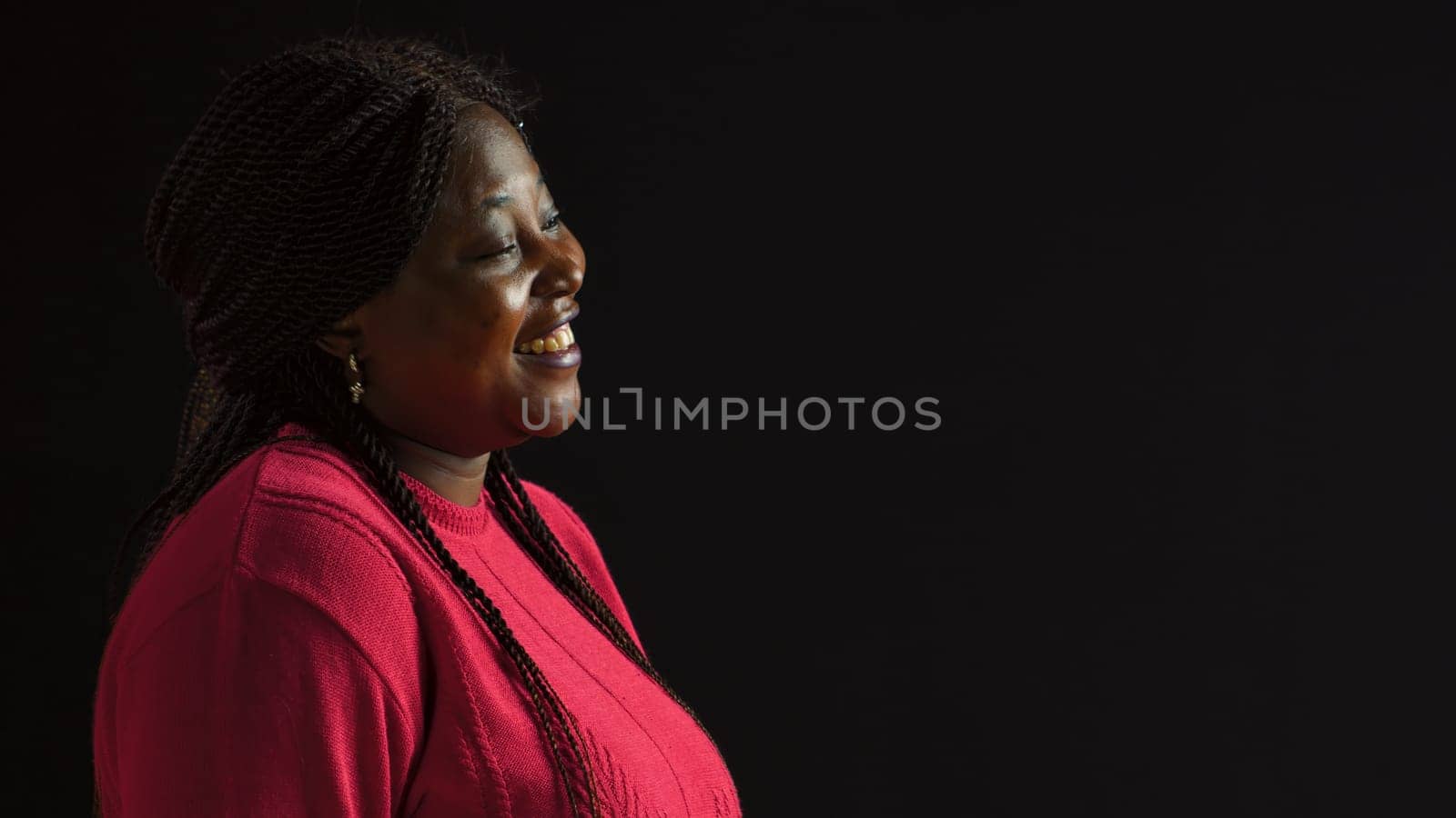 Beautiful african american woman giggling during picture shoot while dressed stylishly in pink. Laughing vibrant female fashionista model on isolated black background. Side-view portrait slow-mo.