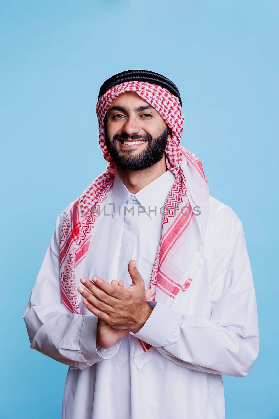 Confident smiling muslim man in traditional robe applauding with hands, expressing support at presentation. Cheerful young arab showing applause gesture and looking at camera