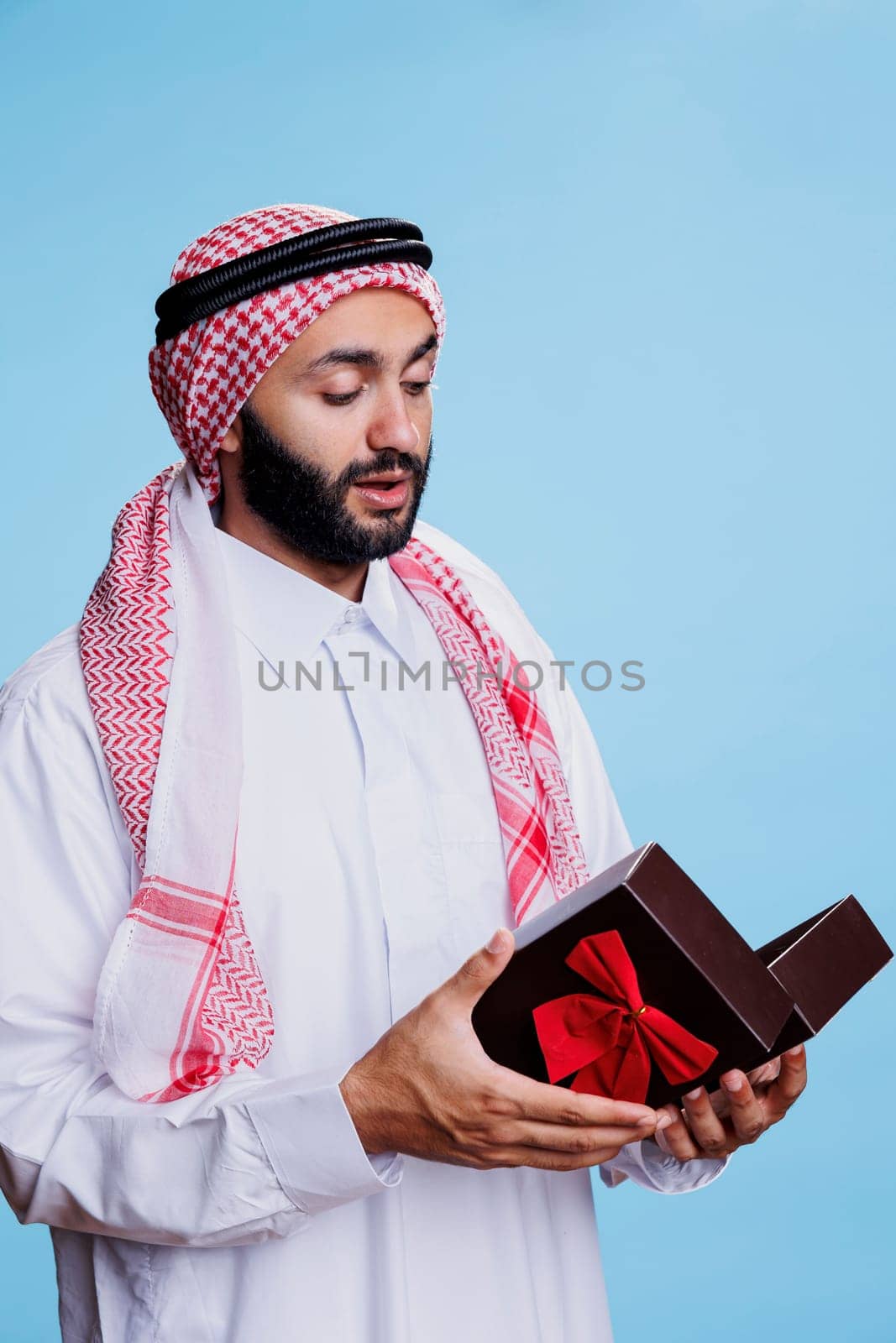 Muslim man wearing traditional attire holding giftbox with bow ribbon and opening festive present. Arab dressed in white thobe standing with giftbox in studio on blue background