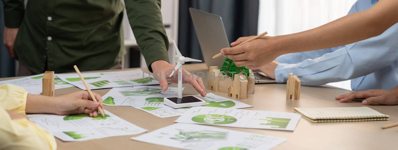 Windmill model represented renewable energy and wooden block represented eco city was placed on green business meeting table with environmental document scatter around. Front view. Delineation.