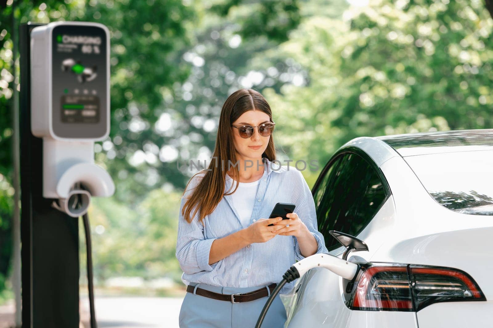 Young woman using smartphone online banking application to pay for electric car battery charging from EV charging station during vacation holiday road trip at national park or summer forest. Exalt