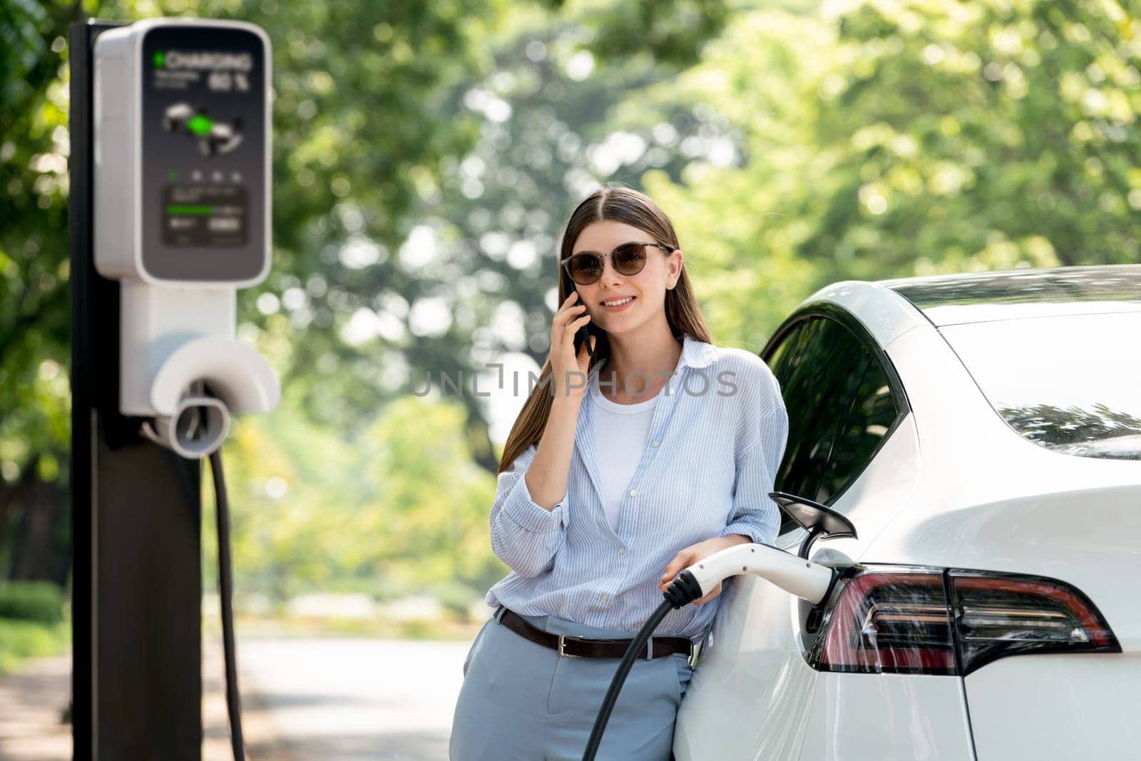 Young woman talking on smartphone while recharging electric car. Exalt by biancoblue