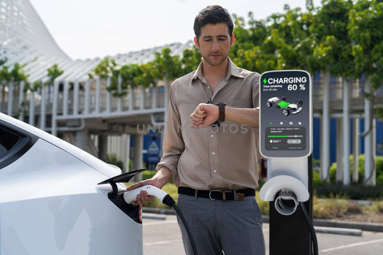 Young man checking time on smartwatch while EV charger to recharging battery from charging station in city mall parking lot. Rechargeable EV car for sustainable eco friendly urban travel. Expedient