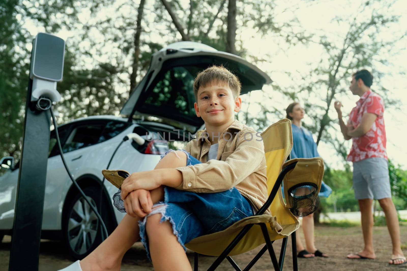 Little boy portrait sitting on camping chair with his family in background. Road trip travel with alternative energy charging station for eco-friendly car concept. Perpetual