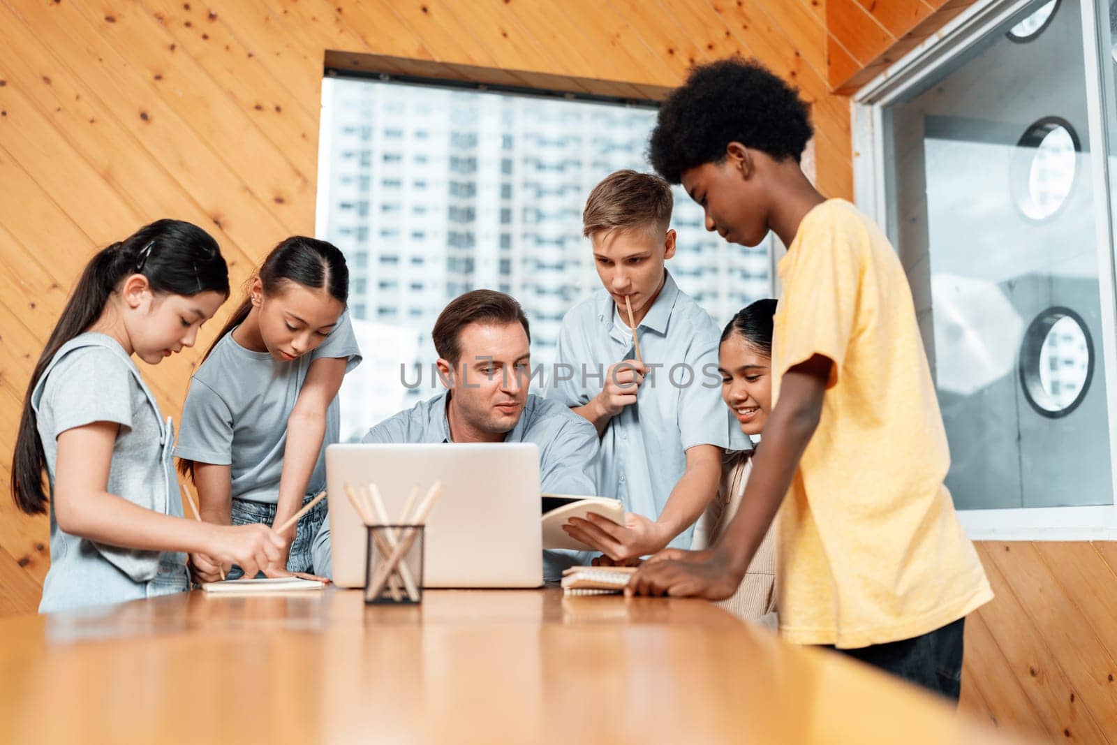 Teacher sitting and giving a lecture on lesson to children in classroom using laptop, talking to children about content in notebook. Students asking about subject instructor teach them. Edification.