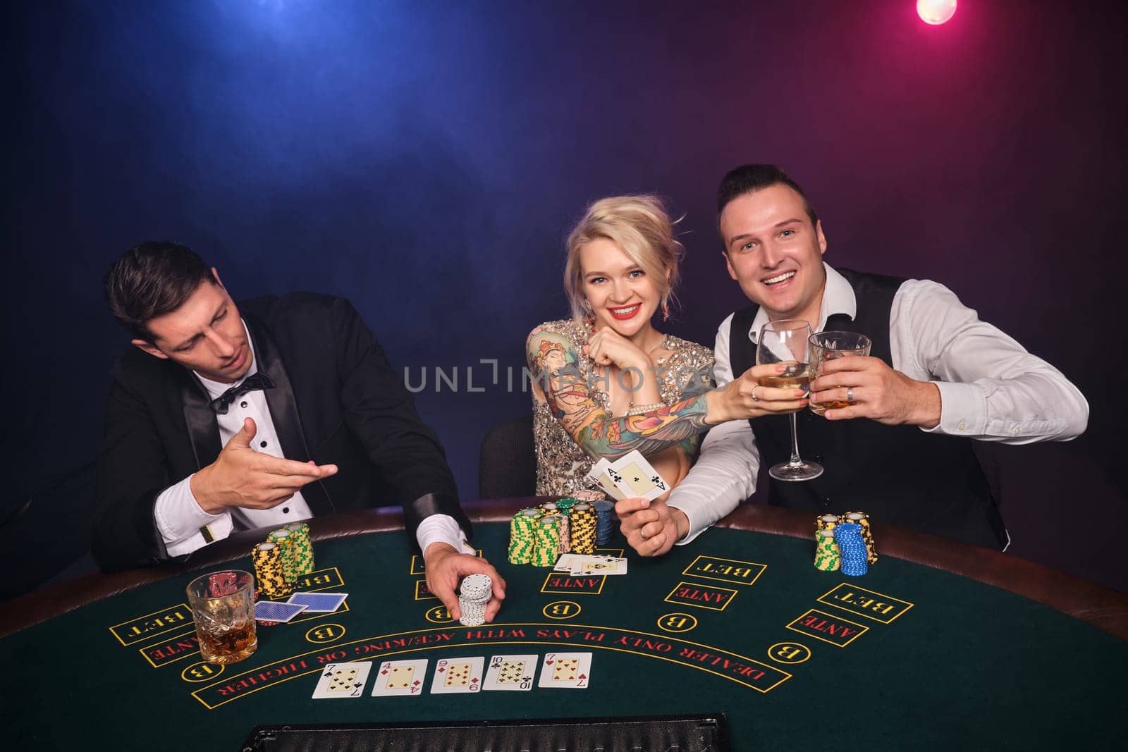 Two elegant male and attractive female are playing poker at casino. Youth are making bets waiting for a big win. They are smiling and posing sitting at the table against a red and blue backlights on black smoke background. Cards, chips, money, gambling, entertainment concept.