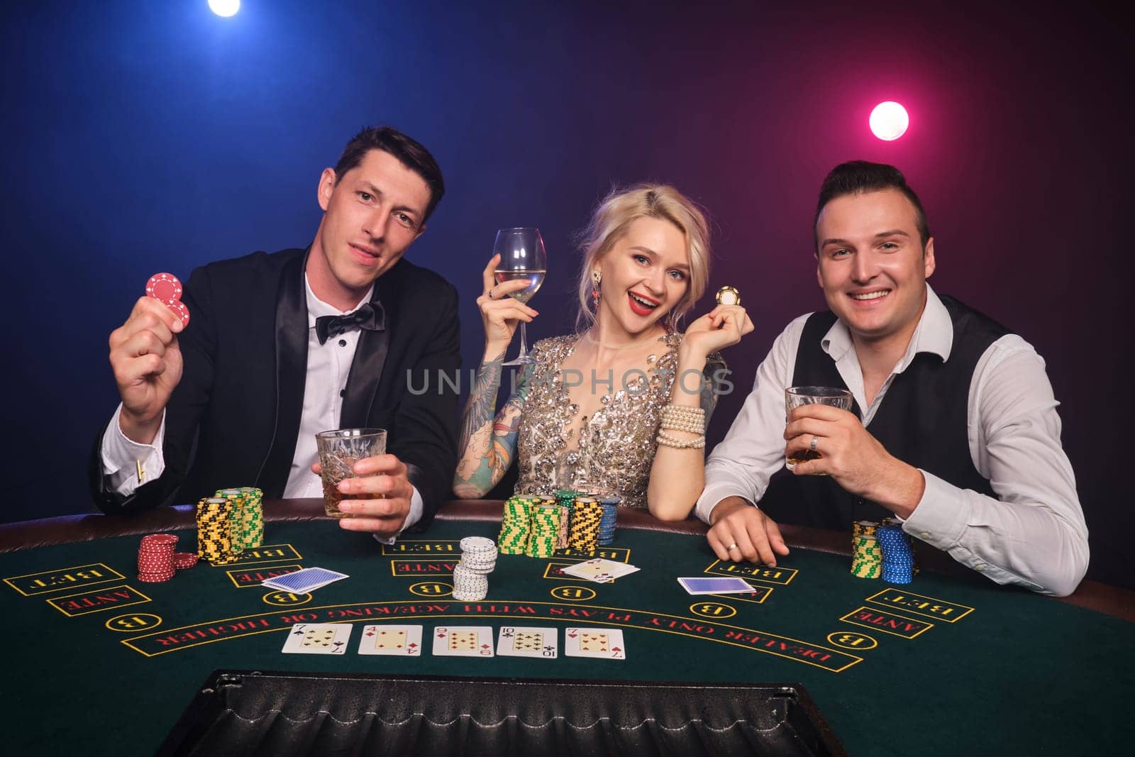 Two good-looking men and amazing maiden are playing poker at casino. Youth are making bets waiting for a big win. They are smiling and looking at the camera sitting at the table against a red and blue backlights on black smoke background. Cards, chips, money, gambling, entertainment concept.